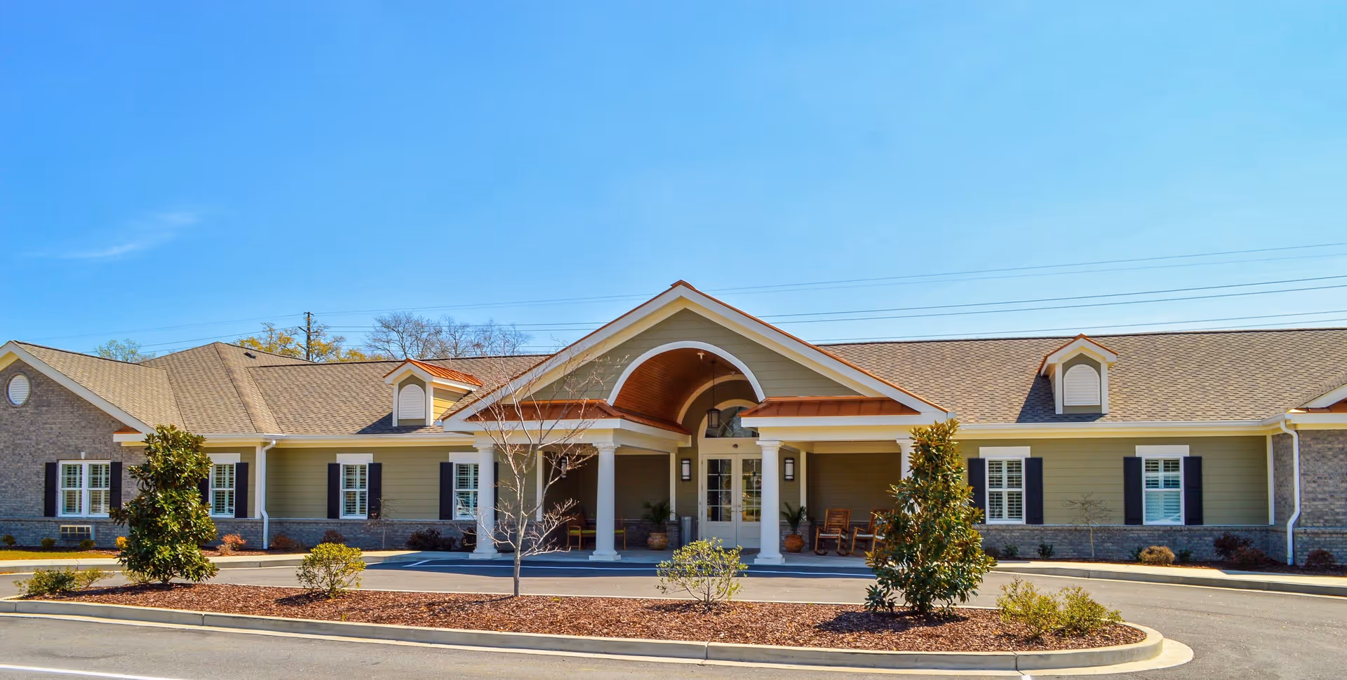Front exterior view of a single-story assisted living and memory care facility with a covered entrance supported by white columns, beige siding, brick accents, and a clear blue sky above. There are small landscaped areas with bushes and trees in front of the building.