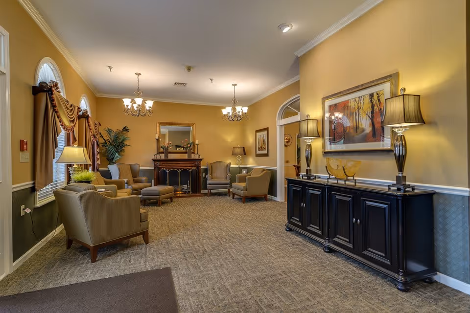 A cozy senior living facility common area with beige walls and carpeted floor. The room features several upholstered armchairs arranged around a fireplace with a decorative mirror above it. Two chandeliers hang from the ceiling, and there are table lamps on a black sideboard and next to the chairs. The windows have elegant drapes, and there is framed artwork on the walls.