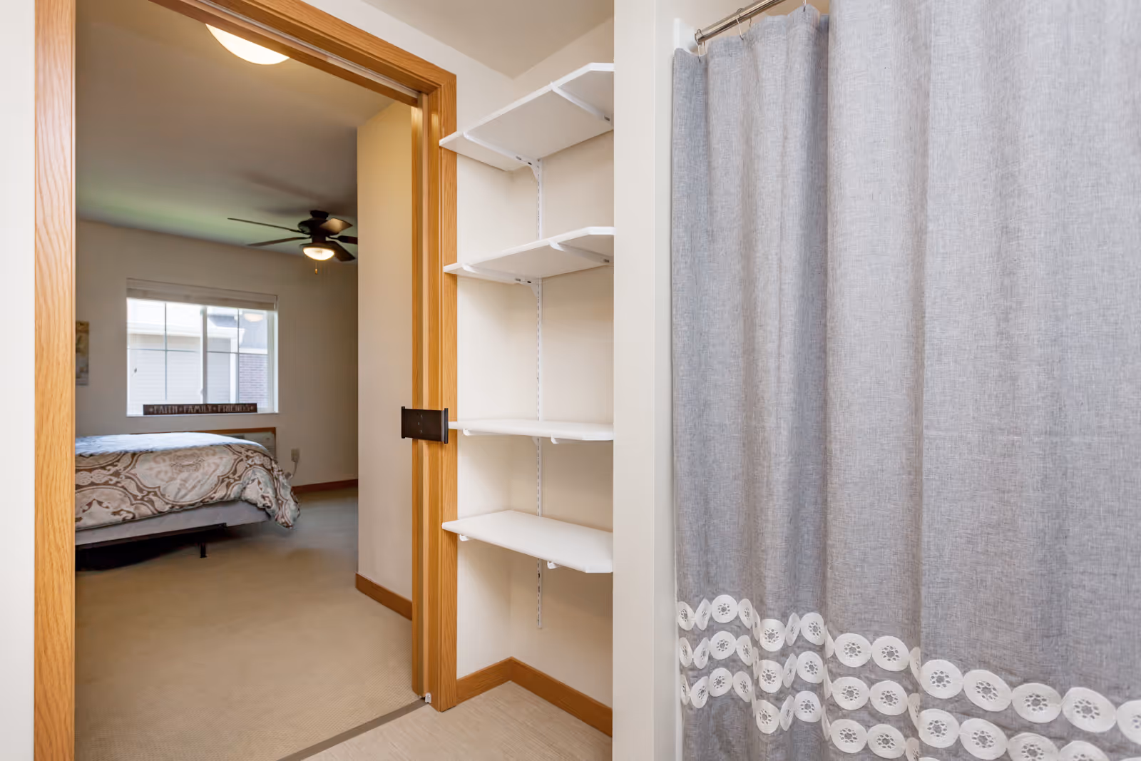 View from a bathroom area showing a gray shower curtain with white decorative trim on the right, white wall-mounted shelves in the center, and an open doorway leading to a bedroom with a bed, window, and ceiling fan in the background.