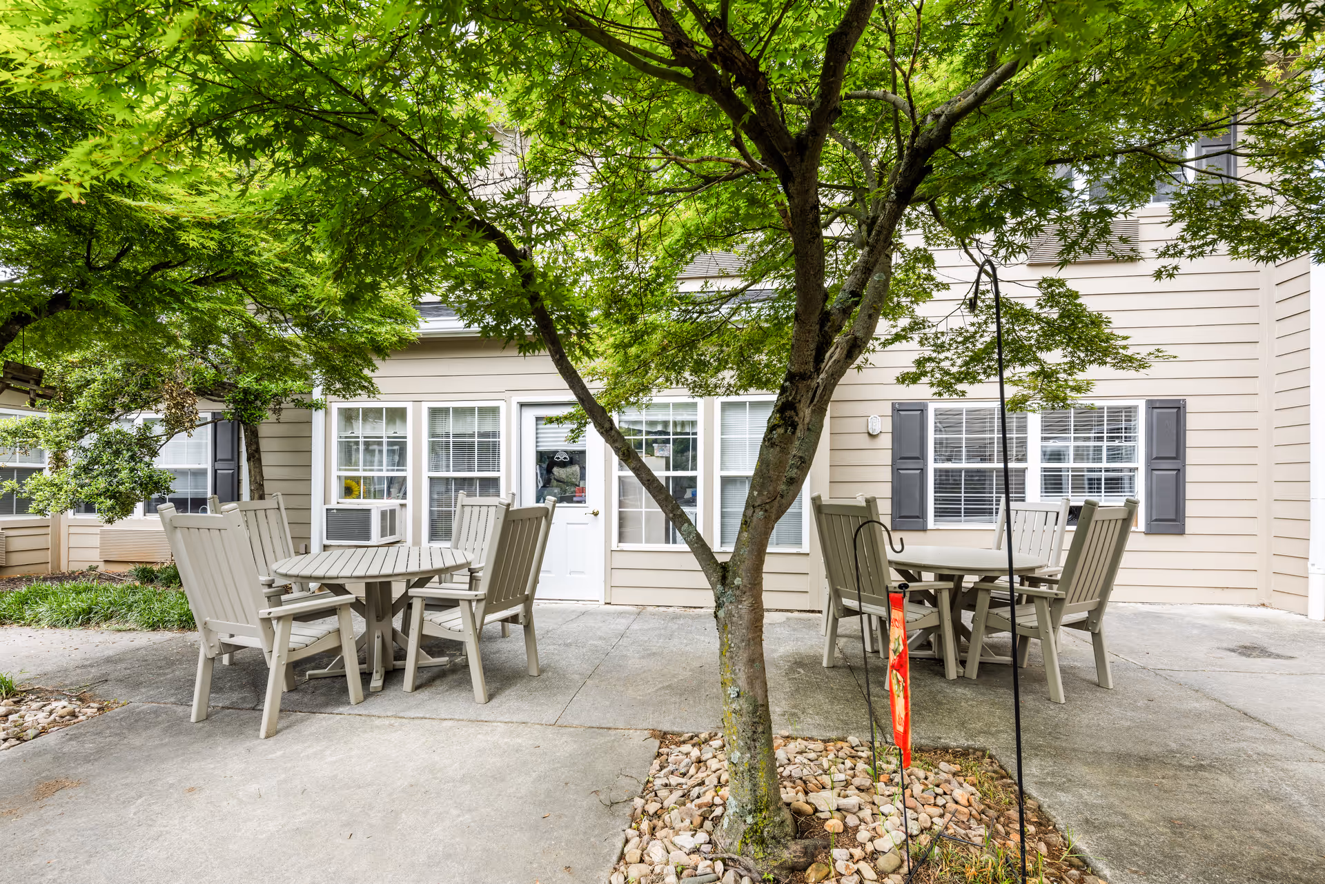 Outdoor patio area with two round tables and several beige chairs under green leafy trees, adjacent to a beige building with white-framed windows and a white door.