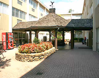 A brick-paved courtyard with a covered wooden gazebo, raised flowerbed, and vending machine between apartment wings.