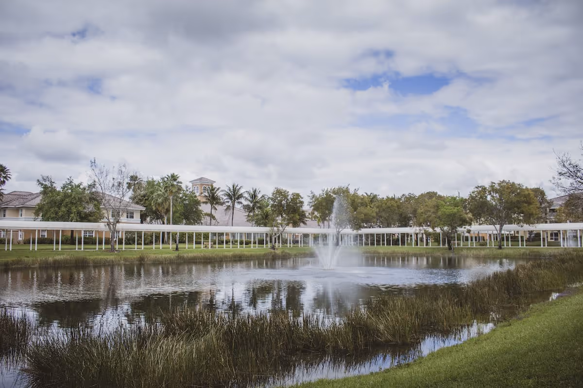 A scenic view of a pond with a water fountain in the center, surrounded by green grass and tall reeds. In the background, there are trees, palm trees, and a covered walkway with buildings visible behind it under a partly cloudy sky.
