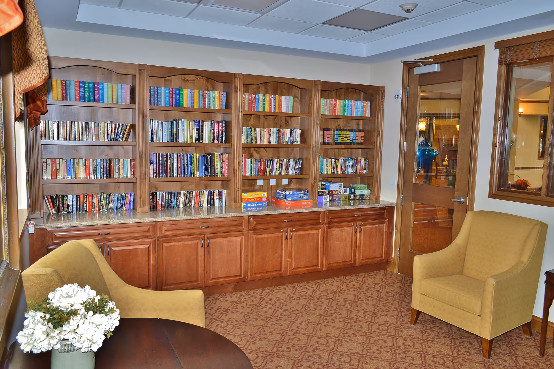 A cozy reading room with wooden bookshelves filled with books and board games, two yellow upholstered armchairs, a small round table with a vase of white flowers, and a wooden door with a window leading to another room.