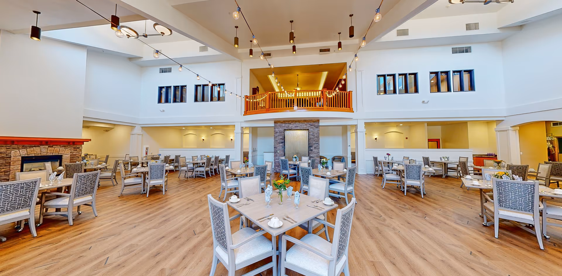A spacious dining room with multiple tables and chairs arranged neatly. The room features high ceilings with hanging light fixtures and large windows near the ceiling allowing natural light. There is a stone fireplace on the left side and a balcony area above the central part of the room. The floor is wooden, and the tables are set with cups, napkins, and small flower arrangements.