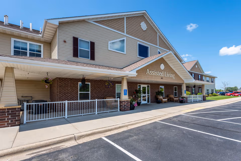 Exterior view of the Edgewood Sartell assisted living facility showing the main entrance with a covered porch, seating area, hanging flower baskets, and a parking lot in front under a clear blue sky.