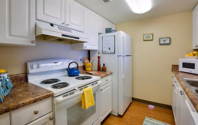 A compact kitchen with white cabinets, a white electric stove with a yellow towel hanging on the handle, a blue kettle on the stove, a white refrigerator with a small whiteboard attached, a microwave on the right countertop, and various kitchen utensils and decorations on the counters. The floor is wooden and there are two small framed pictures on the wall.