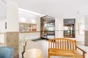 Interior view of a senior living facility lobby area with a wooden bench, a small round table with a plant, and a reception window. The space is well-lit with white walls and large windows near the entrance door.