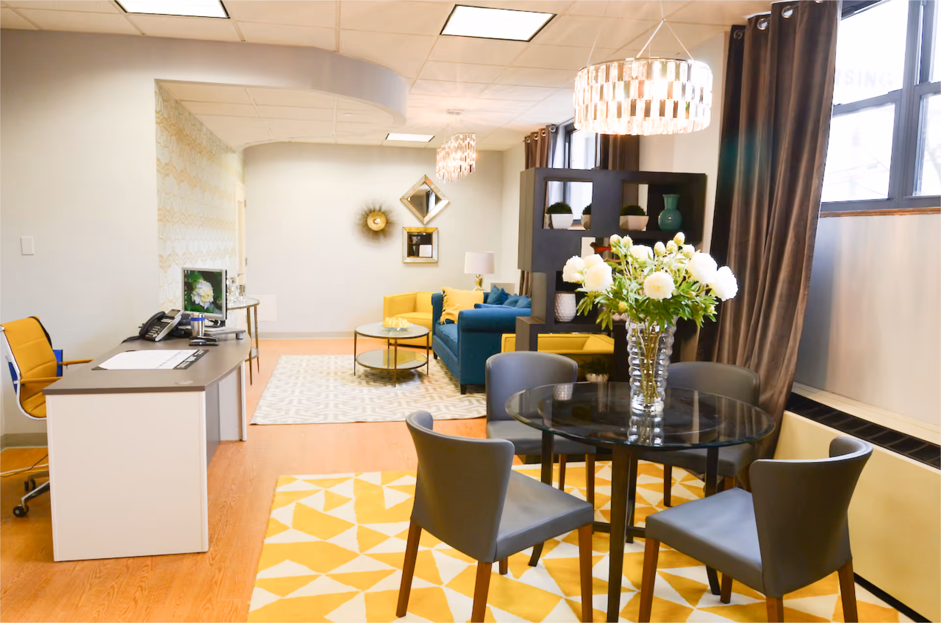 A bright and modern interior space featuring a small round glass dining table with four gray chairs on a yellow and white geometric rug. Behind the table is a black shelving unit with decorative items and a blue couch with yellow cushions. To the left, there is a reception desk with a yellow office chair and a computer monitor. The room has wood flooring, large windows with brown curtains, and two hanging light fixtures.