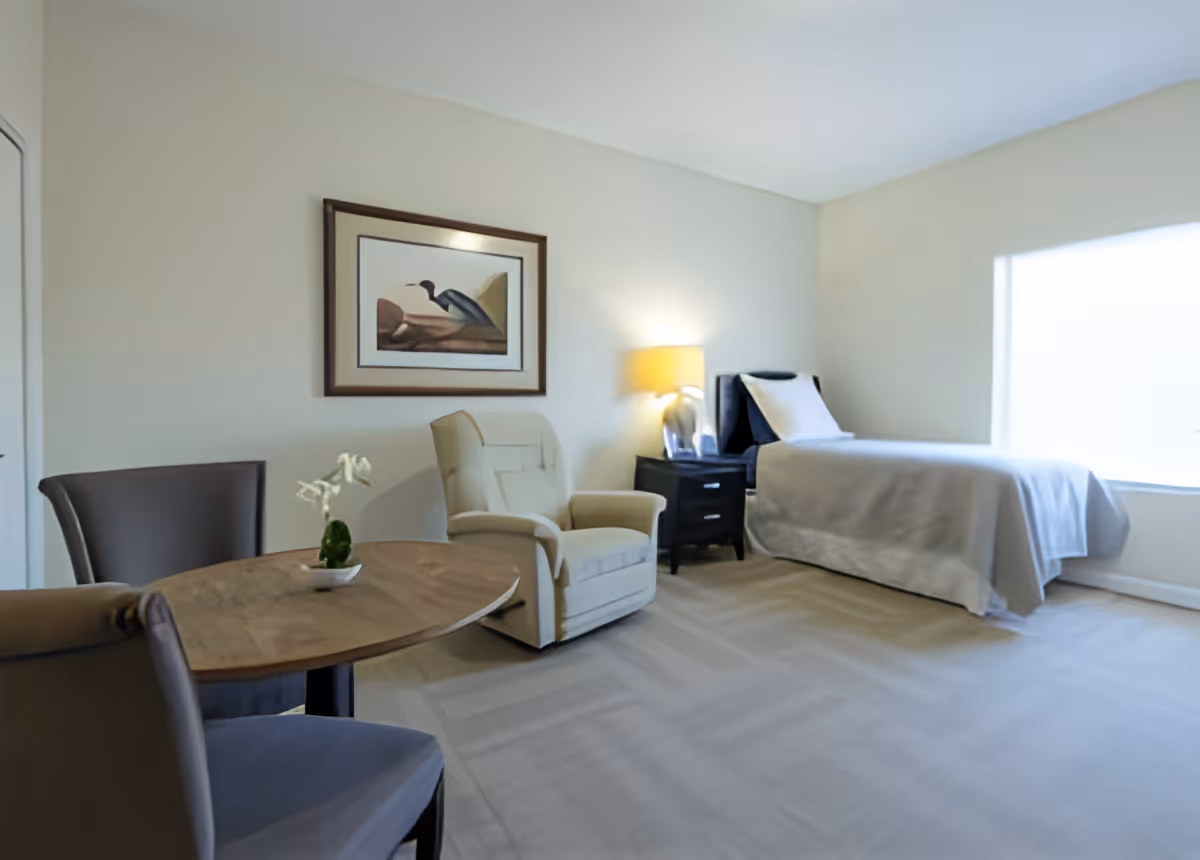 A bright, neatly arranged bedroom with a single bed covered in light gray bedding, a black nightstand with a lamp, a beige armchair, and a small round wooden table with two chairs. A framed picture of a bird hangs on the wall above the armchair.