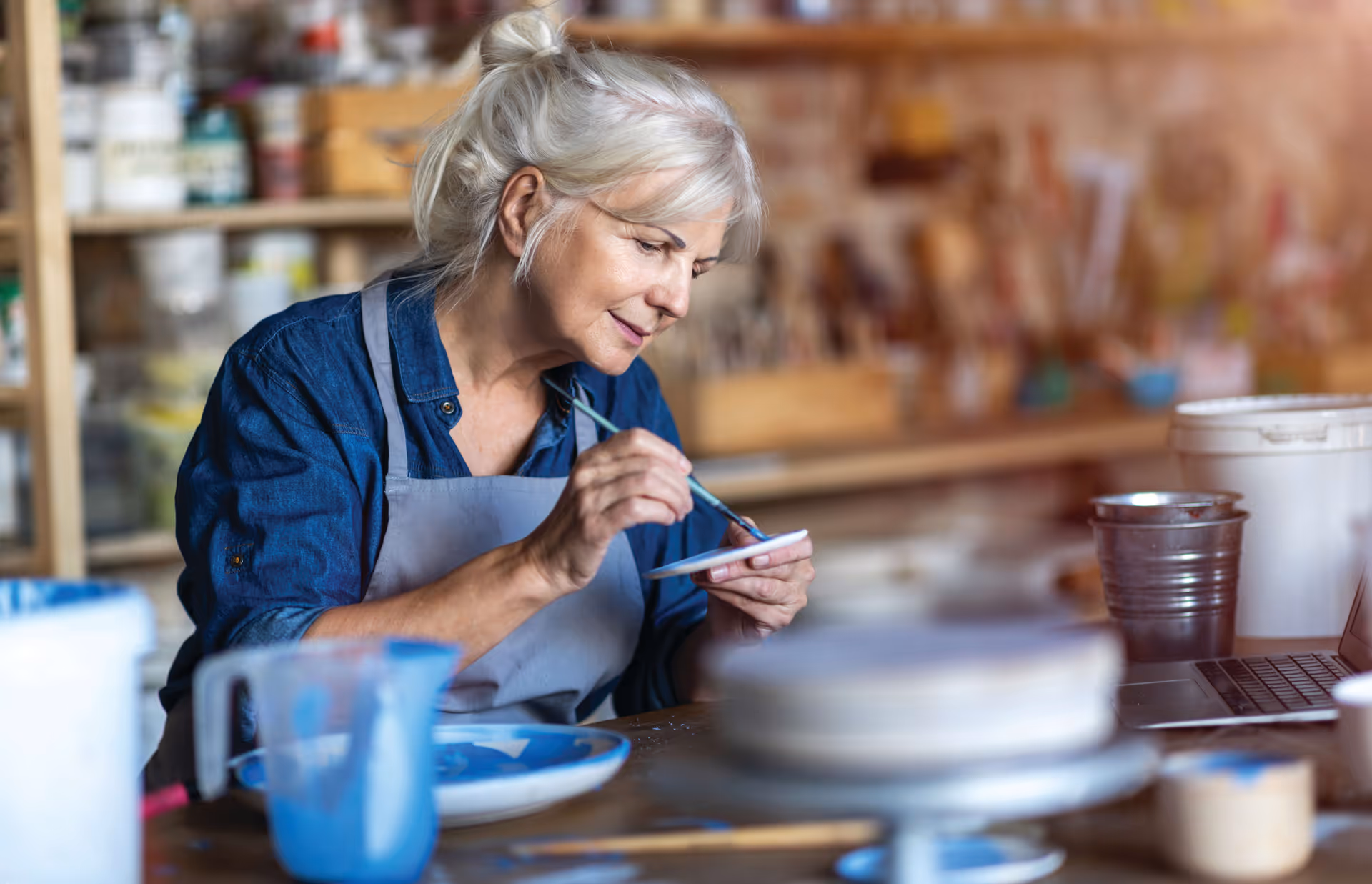 An elderly woman with gray hair tied back is painting a small ceramic plate in a workshop filled with art supplies and pottery. She is wearing a blue shirt and a gray apron, concentrating on her work.