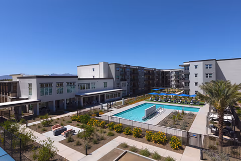 Outdoor view of Mera Goodyear senior living facility featuring a large swimming pool surrounded by lounge chairs with blue umbrellas, landscaped garden areas with pathways, and multi-story residential buildings under a clear blue sky.