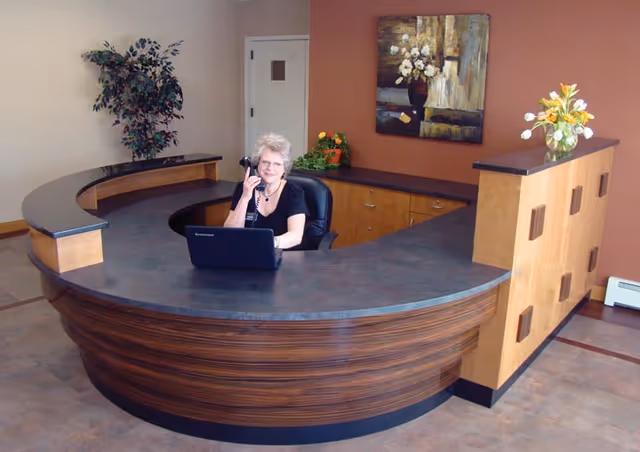 A woman sitting behind a large curved reception desk, talking on a phone with a laptop in front of her. The room has a plant in the corner, a painting on the wall, and a vase with flowers on the desk.
