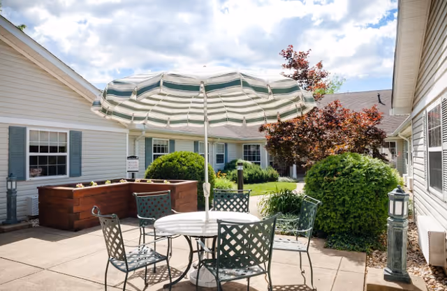 Outdoor patio area with a round metal table and four matching chairs under a large green and white striped umbrella. Surrounding the patio are bushes, a small tree with red leaves, raised garden beds, and the exterior walls of a single-story building with white siding and blue shutters. The sky is partly cloudy.
