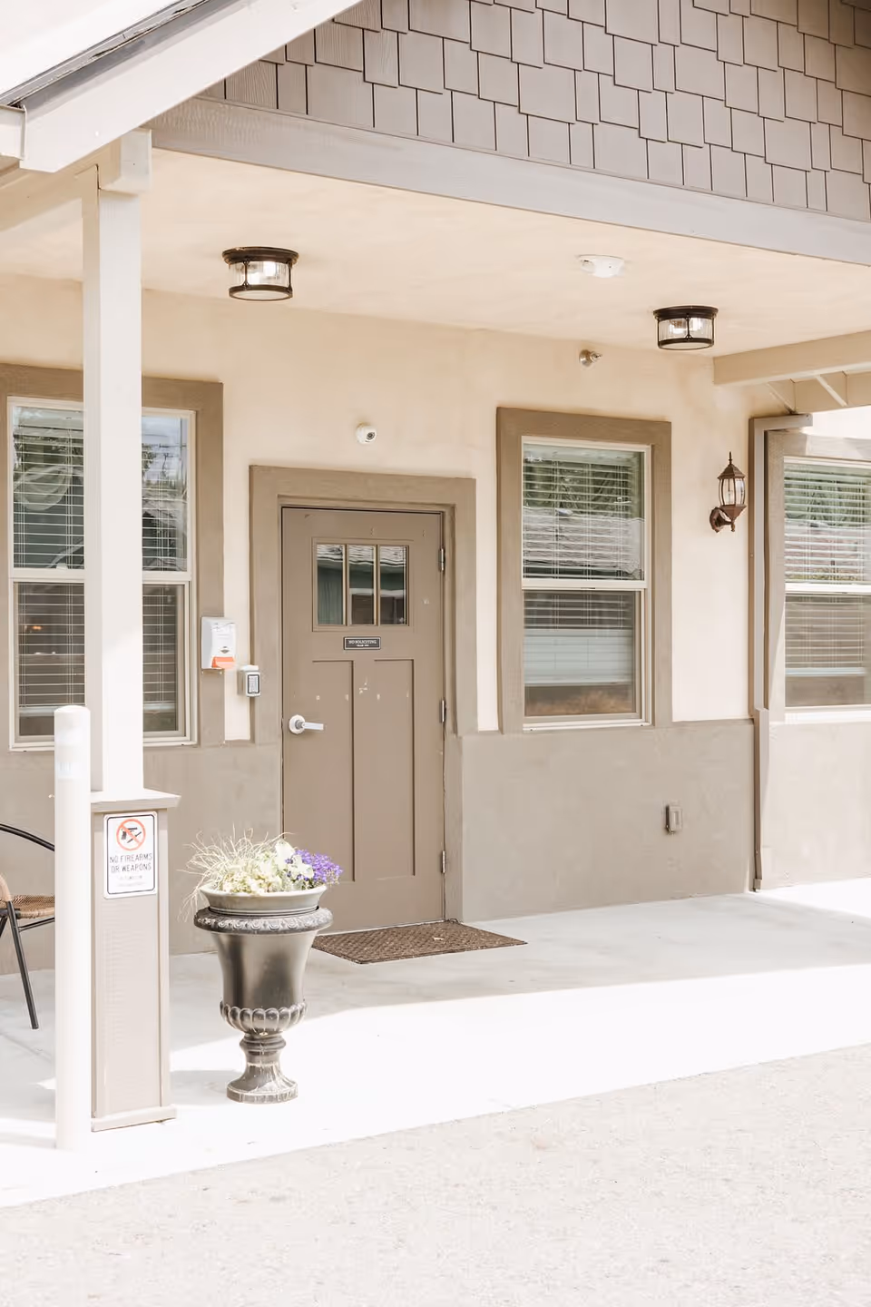 Covered entrance area of a building with a brown door, two windows with blinds, a decorative black planter with flowers, and a sign on a pillar stating no firearms or weapons allowed.