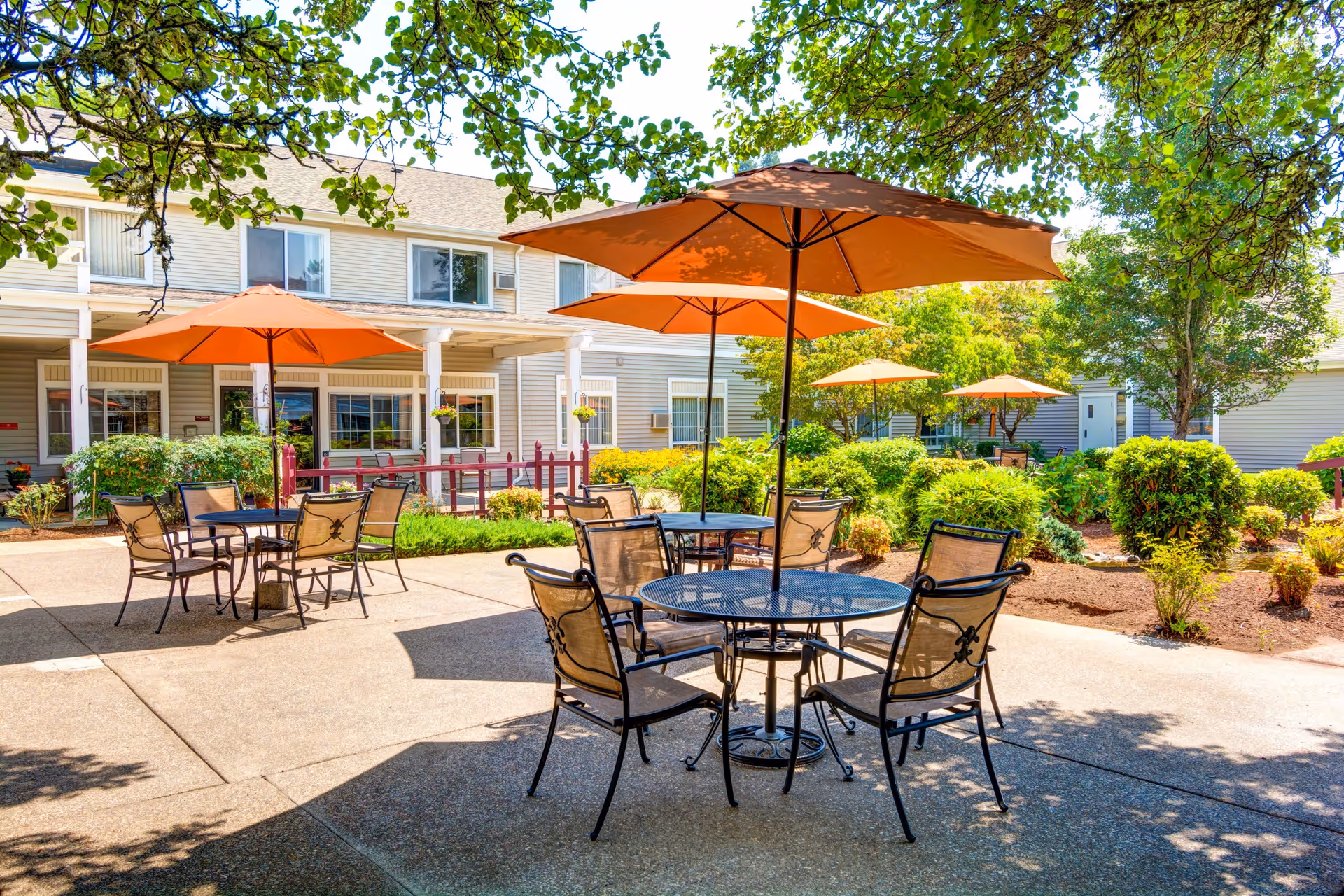 Outdoor patio area at The Oaks at Lebanon featuring multiple round metal tables with orange umbrellas and chairs, surrounded by green bushes and trees, with a two-story building in the background.