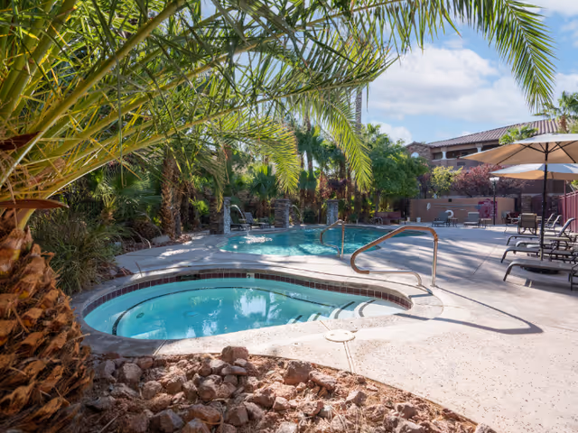 Outdoor pool area at a senior living facility featuring a hot tub in the foreground, a larger swimming pool in the background, surrounded by palm trees and lounge chairs with umbrellas under a clear blue sky.