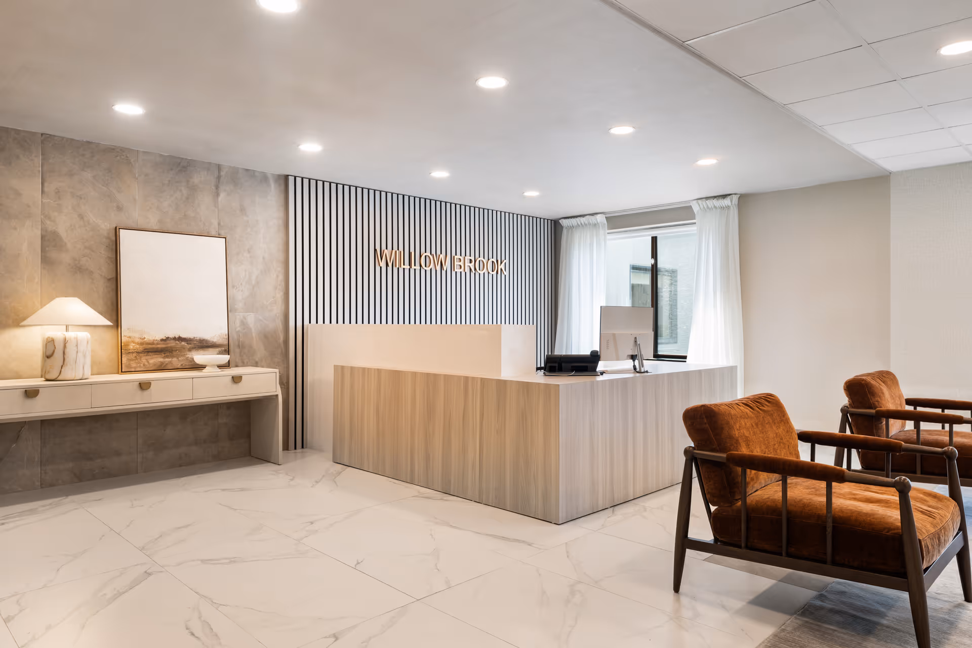 Reception area of Willow Brook Rehabilitation and Healthcare Center featuring a modern wooden reception desk, two brown cushioned chairs with wooden frames, a console table with a lamp and artwork, and a window with white curtains.