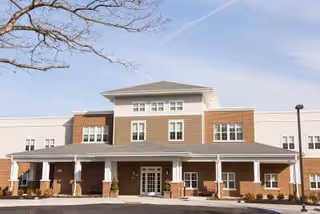 Front exterior view of a senior living facility building with a covered entrance, multiple windows, and a clear sky above.
