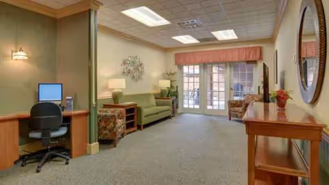A spacious senior living lounge with couches, seating, lamps, and glass doors at the far end.