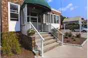 Entrance to Eastgate Nursing & Rehabilitation Center showing a small set of stairs with white railings leading to a glass door under a green awning. There is a sign on the right side with the facility name, some landscaping with plants, and a clear blue sky above.