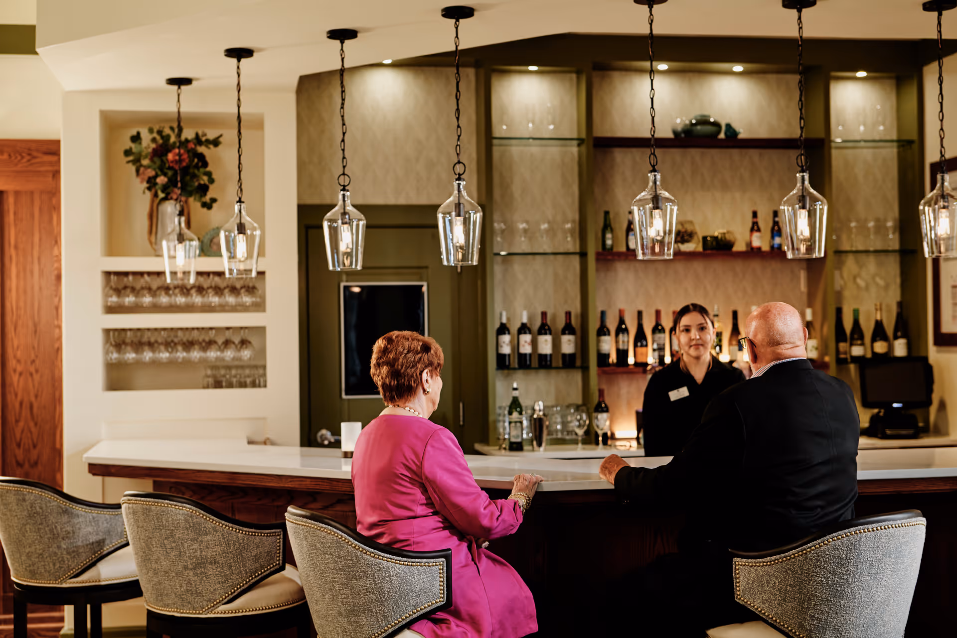 Two older adults sit at a bar talking with a staff member, with wine bottles on shelves and pendant lights overhead.