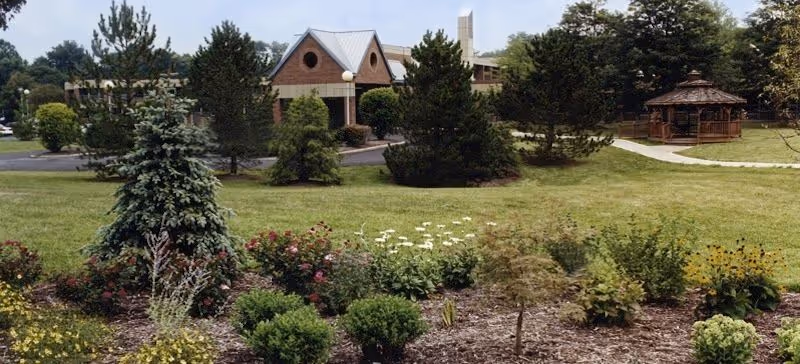 Landscaped lawn and garden beds with trees, a gazebo, and the front entrance of a brick community building.