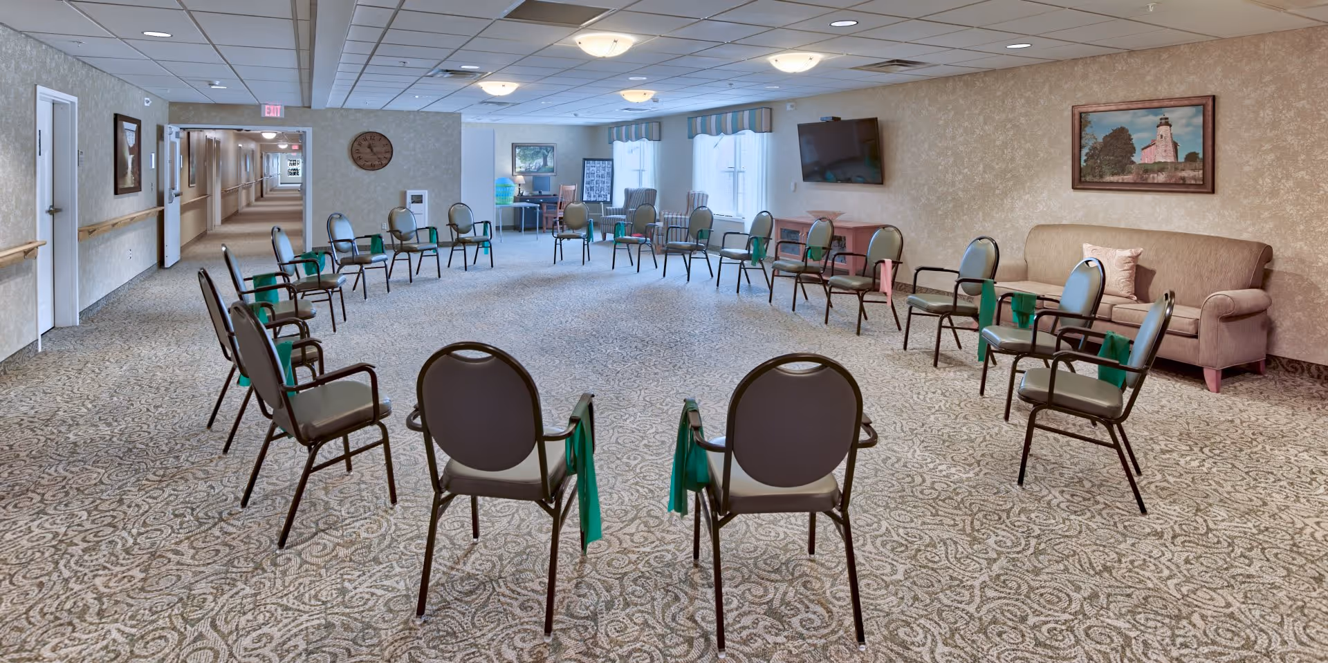 Spacious assisted-living activity room with chairs arranged in a large circle, a sofa and wall-mounted TV, and windows at the far end.