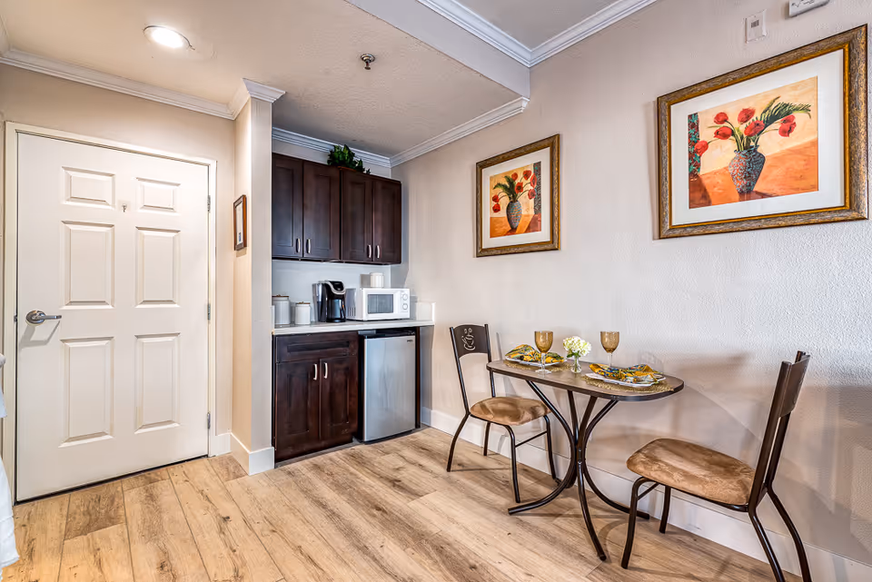 Small dining area with a table set for two, featuring two chairs with cushioned seats, two plates with napkins, two glasses, and a small flower vase. Adjacent to the dining area is a kitchenette with dark wood cabinets, a microwave, a coffee maker, and a mini refrigerator. The walls are light-colored with two framed floral paintings. The floor is wood, and there is a white door on the left side of the image.