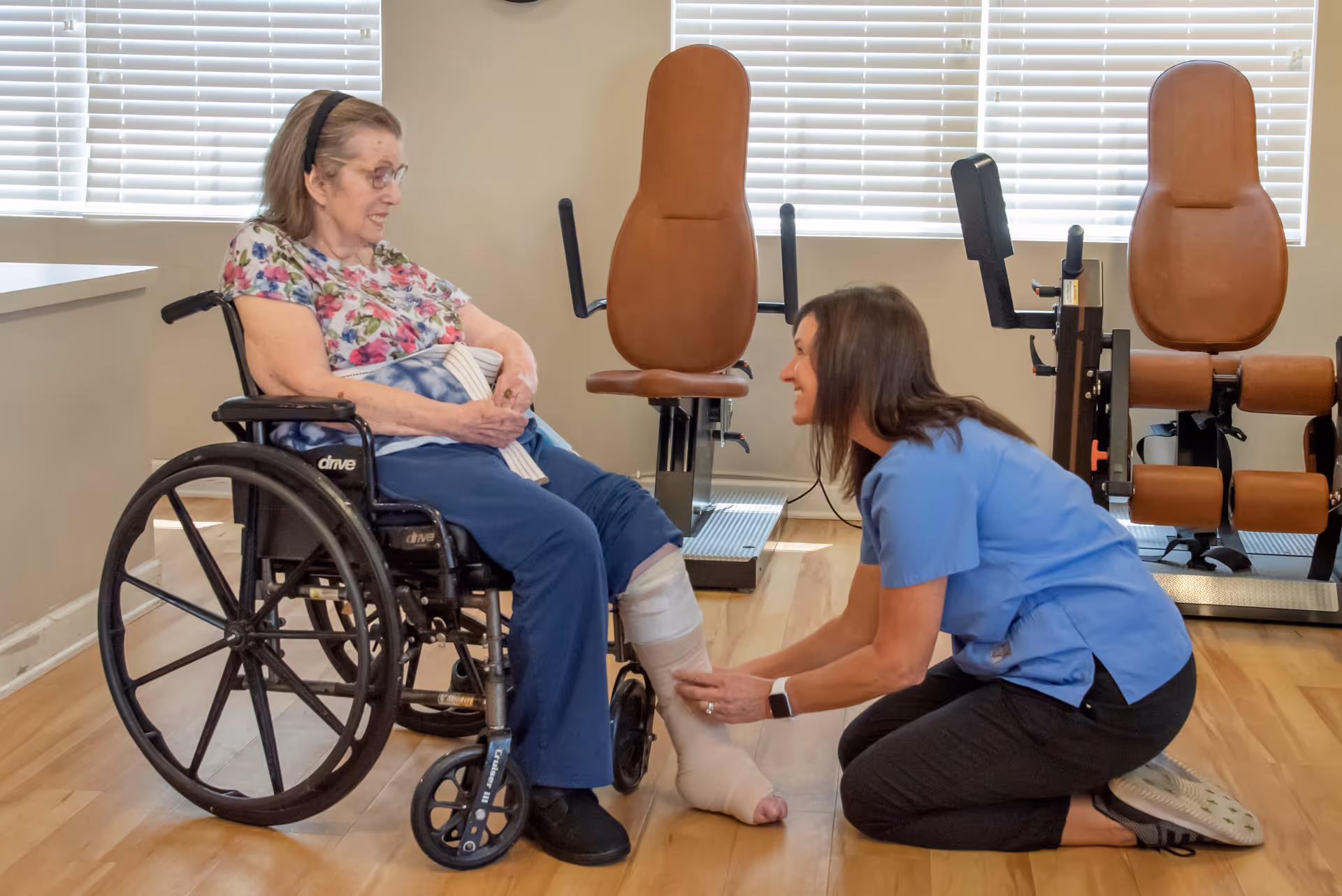 A healthcare worker in blue scrubs kneels on the floor, smiling and attending to an elderly woman seated in a wheelchair with a bandaged leg. The setting appears to be a rehabilitation or therapy room with exercise equipment and large windows with blinds in the background.