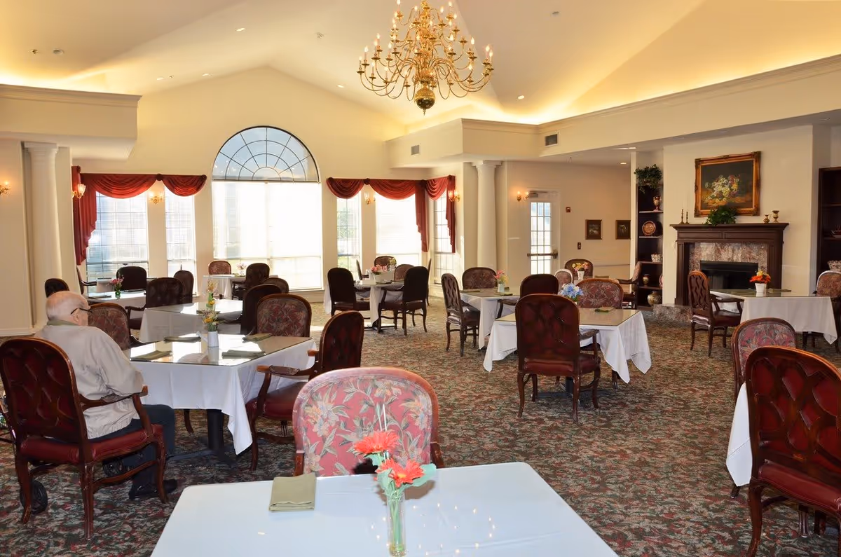 A spacious dining room with multiple tables covered in white tablecloths, each set with floral centerpieces. The room has large windows with red curtains, a high ceiling with a large chandelier, and a fireplace with a painting above it. An elderly man is seated at one of the tables.