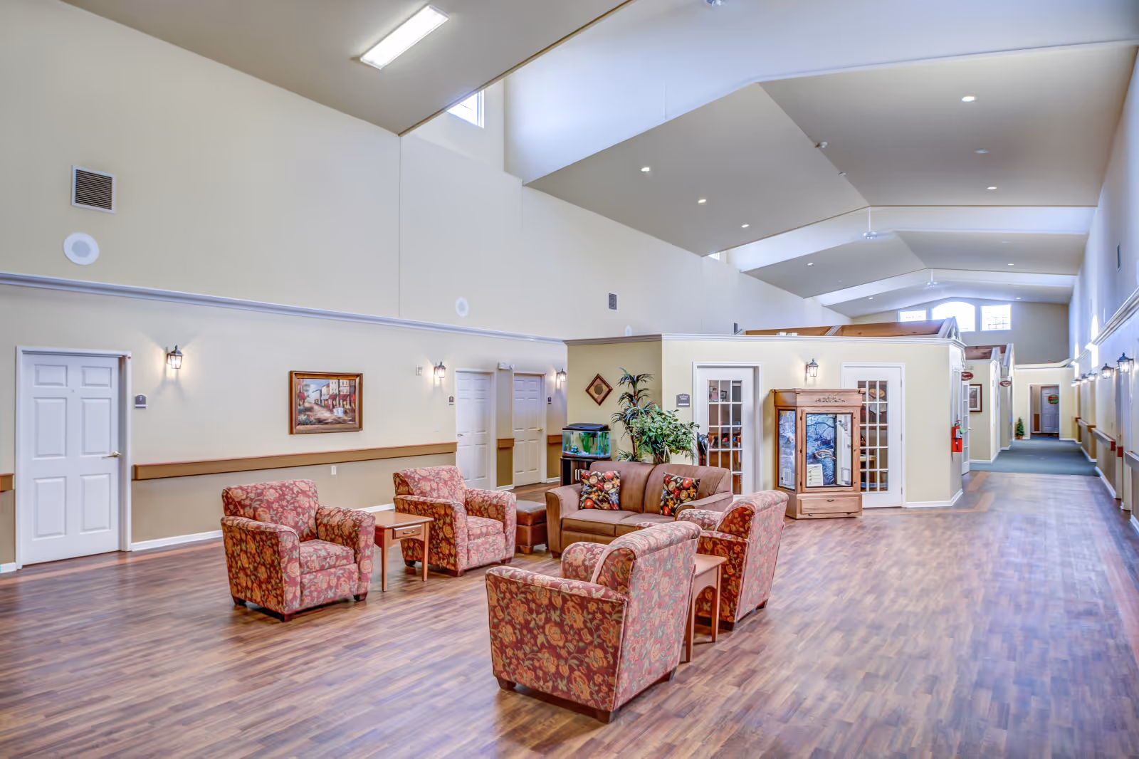 Spacious communal lounge in an assisted living facility with upholstered chairs and sofas arranged on wood flooring under high vaulted ceilings.