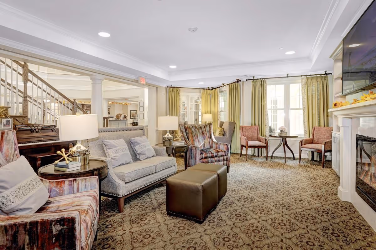 A bright and spacious living room area in a senior living facility with patterned carpet, multiple upholstered chairs and sofas, side tables with lamps, a piano, large windows with green curtains, and a wall-mounted TV above a white fireplace.