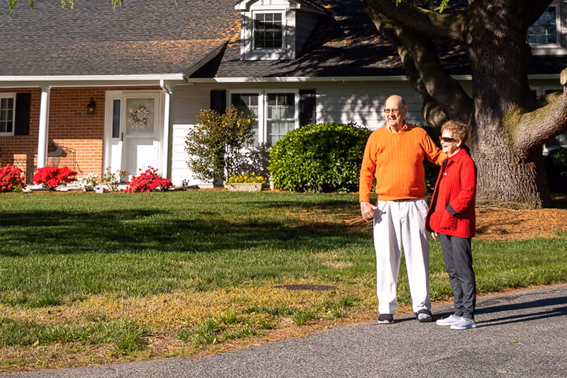 An elderly couple standing outside on a paved path in front of a house with a well-maintained lawn, bushes, and flowers. The man is wearing a bright orange sweater and white pants, and the woman is wearing a red jacket and dark pants. They are standing near a large tree and appear to be enjoying a sunny day.