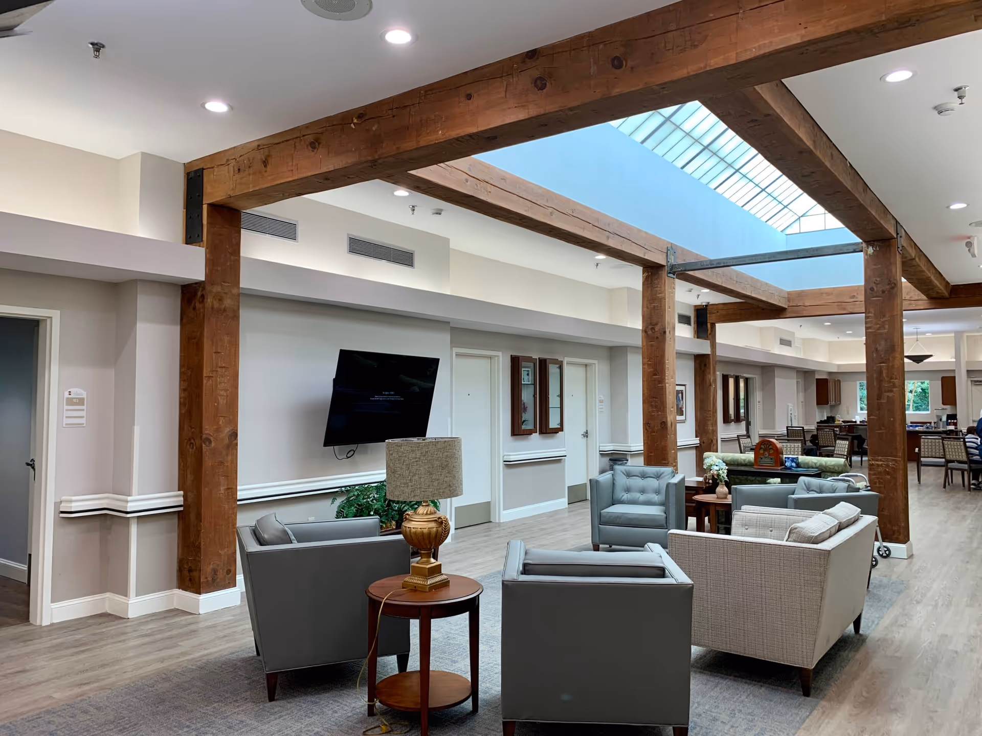 A spacious lobby area featuring modern seating arrangements, wooden beams, and a large skylight, with a television mounted on the wall and a small table with a lamp.