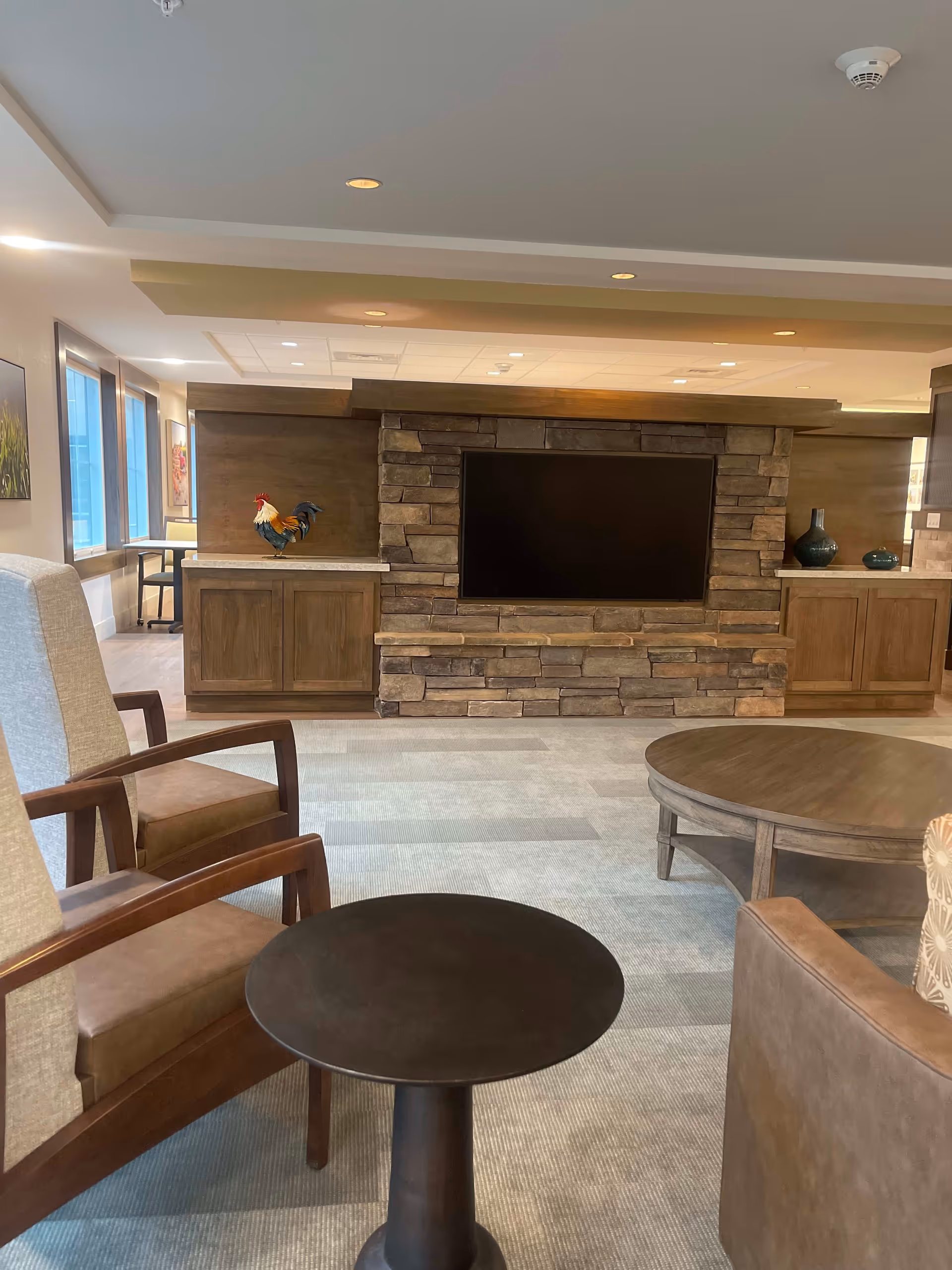 A cozy living room area with a stone fireplace and a large flat-screen TV mounted above it. The room features wooden cabinets on either side of the fireplace, decorated with a colorful rooster figurine and two blue vases. There are comfortable chairs and round wooden tables in the foreground, with large windows letting in natural light on the left side.