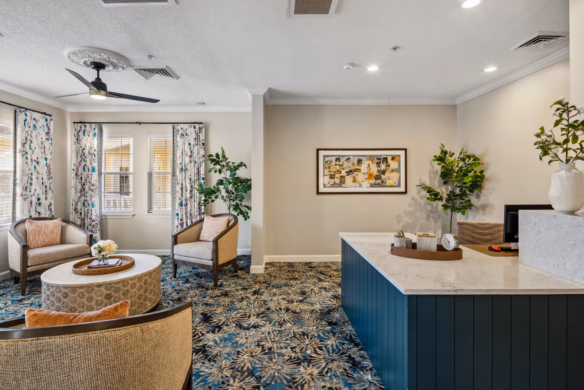 Bright reception lounge with chairs, a round coffee table, potted plants, framed art, and a marble-topped front desk.