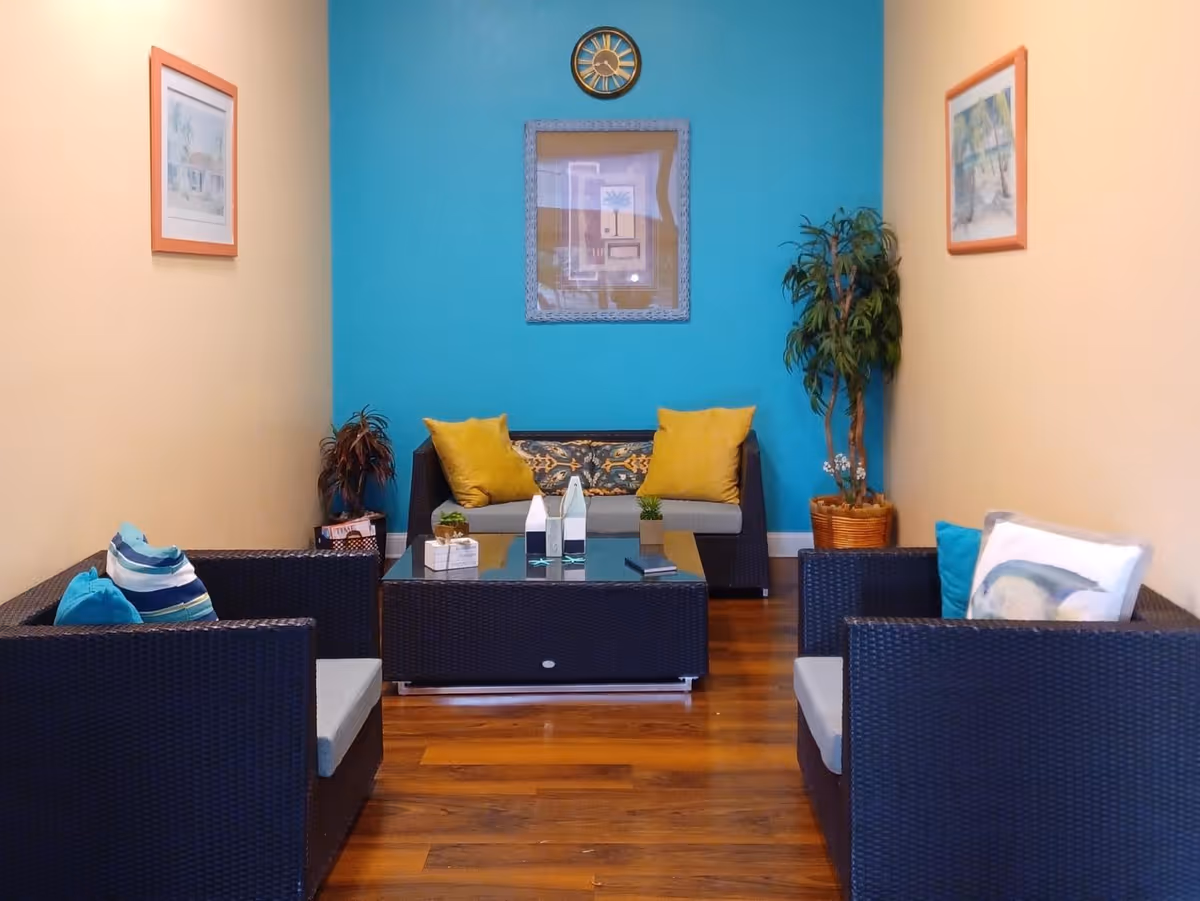 Small communal seating area with wicker sofas and chairs arranged around a glass coffee table against a teal accent wall with artwork and potted plants.