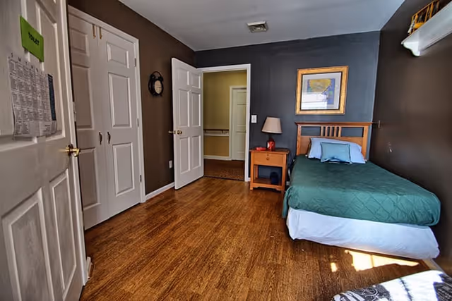 A small bedroom with a single bed covered in a green quilt and a blue pillow. Next to the bed is a wooden nightstand with a lamp and a red object. The walls are painted dark gray, and there is a framed picture hanging above the bed. The floor is wooden, and there are two white doors on the left side of the room, one of which is open, showing a hallway with yellow walls.