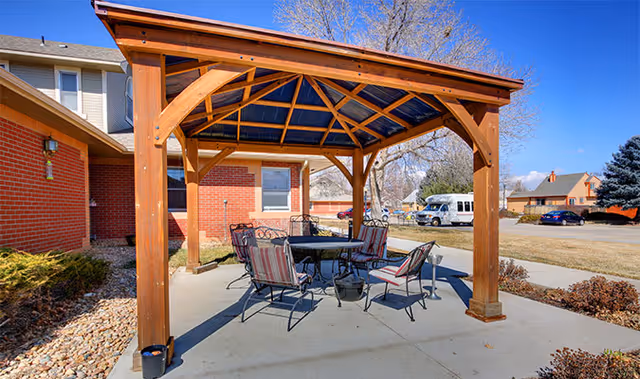Outdoor seating area with a wooden pergola providing shade over a round table surrounded by six metal chairs with cushions. The area is paved with concrete and is adjacent to a red brick building. In the background, there is a parking lot with a white van and other vehicles, along with some trees and houses under a clear blue sky.