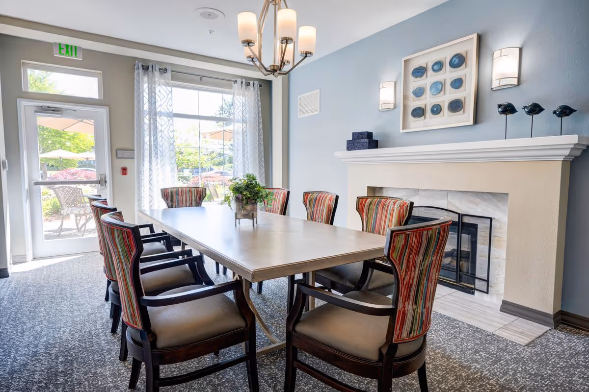 A bright dining room with a rectangular table surrounded by eight upholstered chairs with striped backs. The room features a fireplace with decorative items on the mantel, a large window with sheer curtains letting in natural light, and a chandelier hanging above the table. Outside, patio furniture and umbrellas are visible through the glass door and window.