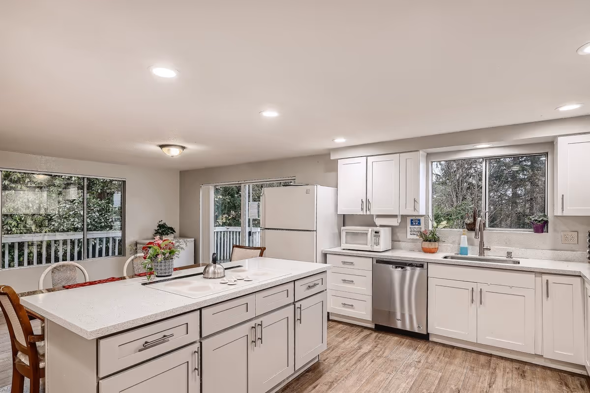 A bright and modern kitchen with white cabinets and drawers, a large island countertop with a built-in stove and a kettle on it. There are two large windows showing greenery outside, a white refrigerator, a microwave, a dishwasher, and a sink with a faucet. The kitchen has wooden flooring and recessed ceiling lights.