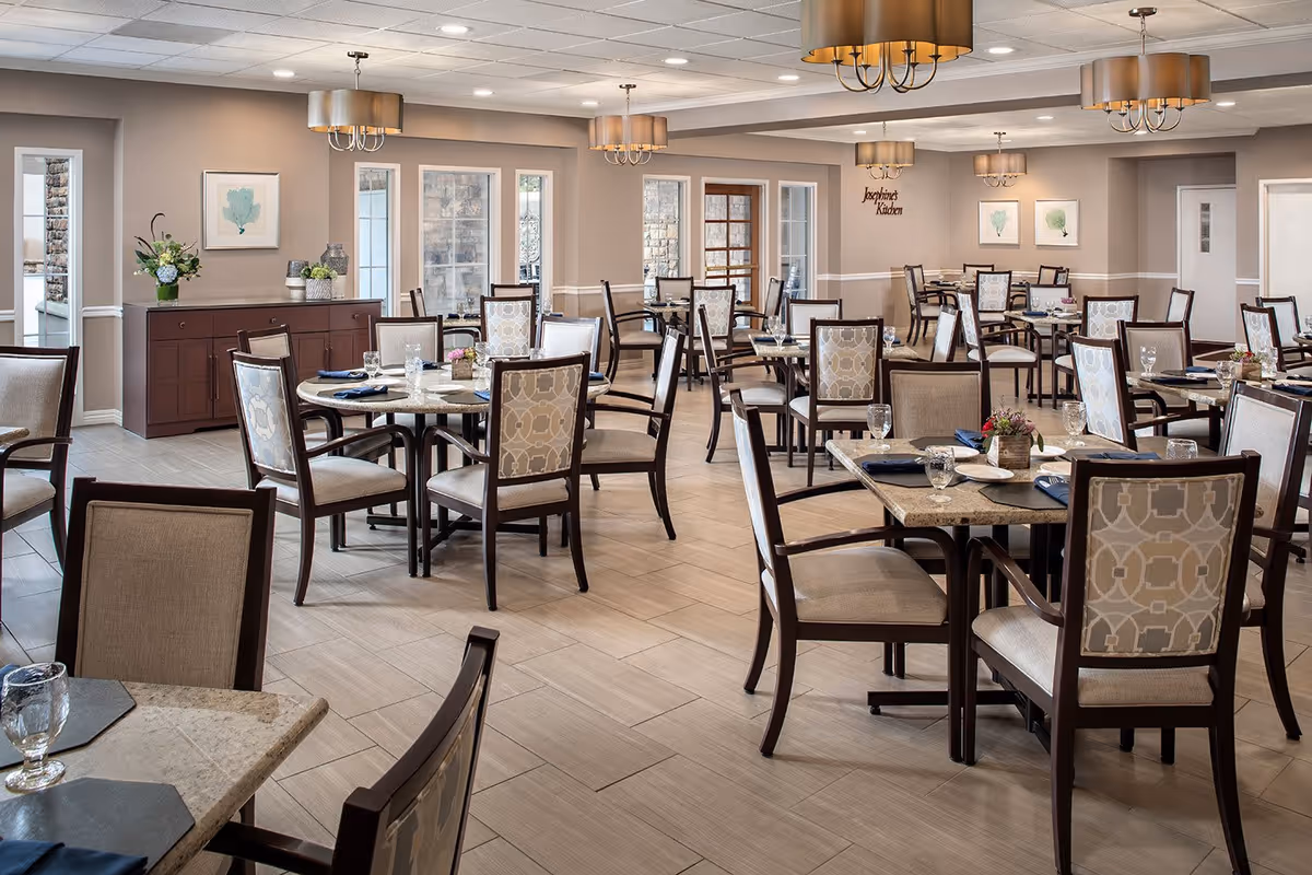 A spacious and well-lit dining room in a senior living facility with multiple tables and chairs arranged neatly. Each table is set with placemats, napkins, glasses, and small floral centerpieces. The room features beige walls, large windows, decorative wall art, and elegant ceiling light fixtures.