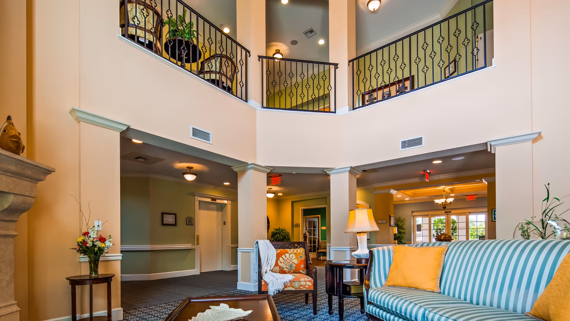 A spacious and well-lit senior living facility common area with a high ceiling and an upper balcony with decorative railings. The room features a striped blue and white sofa with yellow pillows, an orange floral armchair with a white throw, a side table with a lamp, and a small round table with a vase of flowers. The walls are painted in soft beige and green tones, and there is an elevator visible in the background.