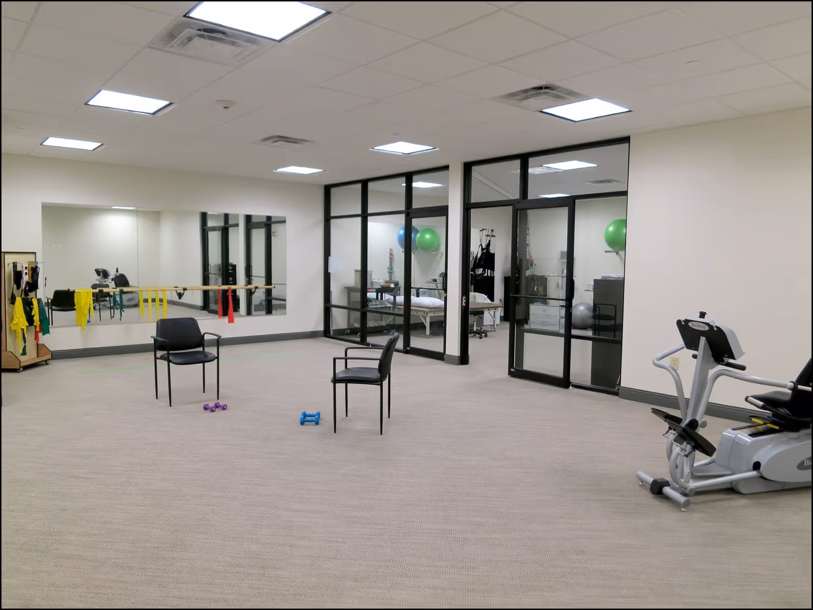 A spacious exercise room with beige carpet and white walls. The room contains two black chairs, small dumbbells on the floor, resistance bands hanging on a ballet barre attached to a mirrored wall, and an exercise bike on the right. Through glass doors and windows, a smaller room with exercise balls and medical equipment is visible.