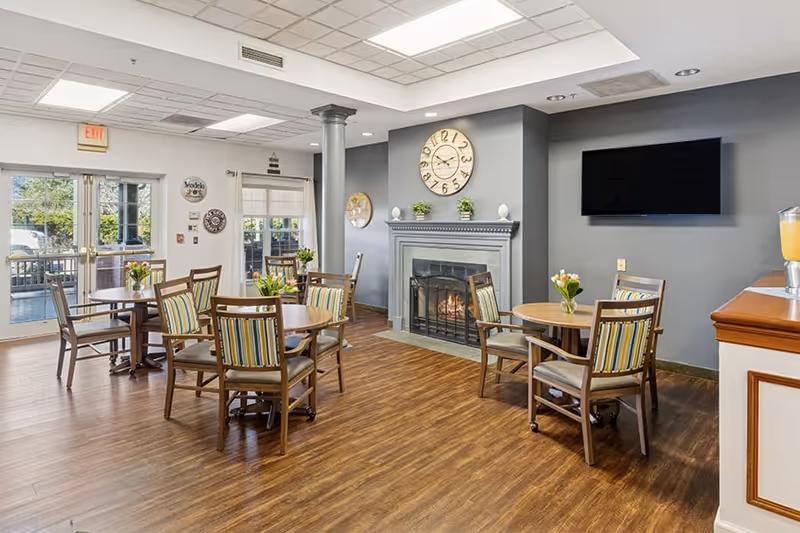 A cozy senior living dining area with several round wooden tables and chairs featuring striped cushions. There is a fireplace with a large clock above it on a gray accent wall, a flat-screen TV mounted on the wall, and glass doors leading to an outdoor patio. The room has wood flooring and a ceiling with recessed lighting.