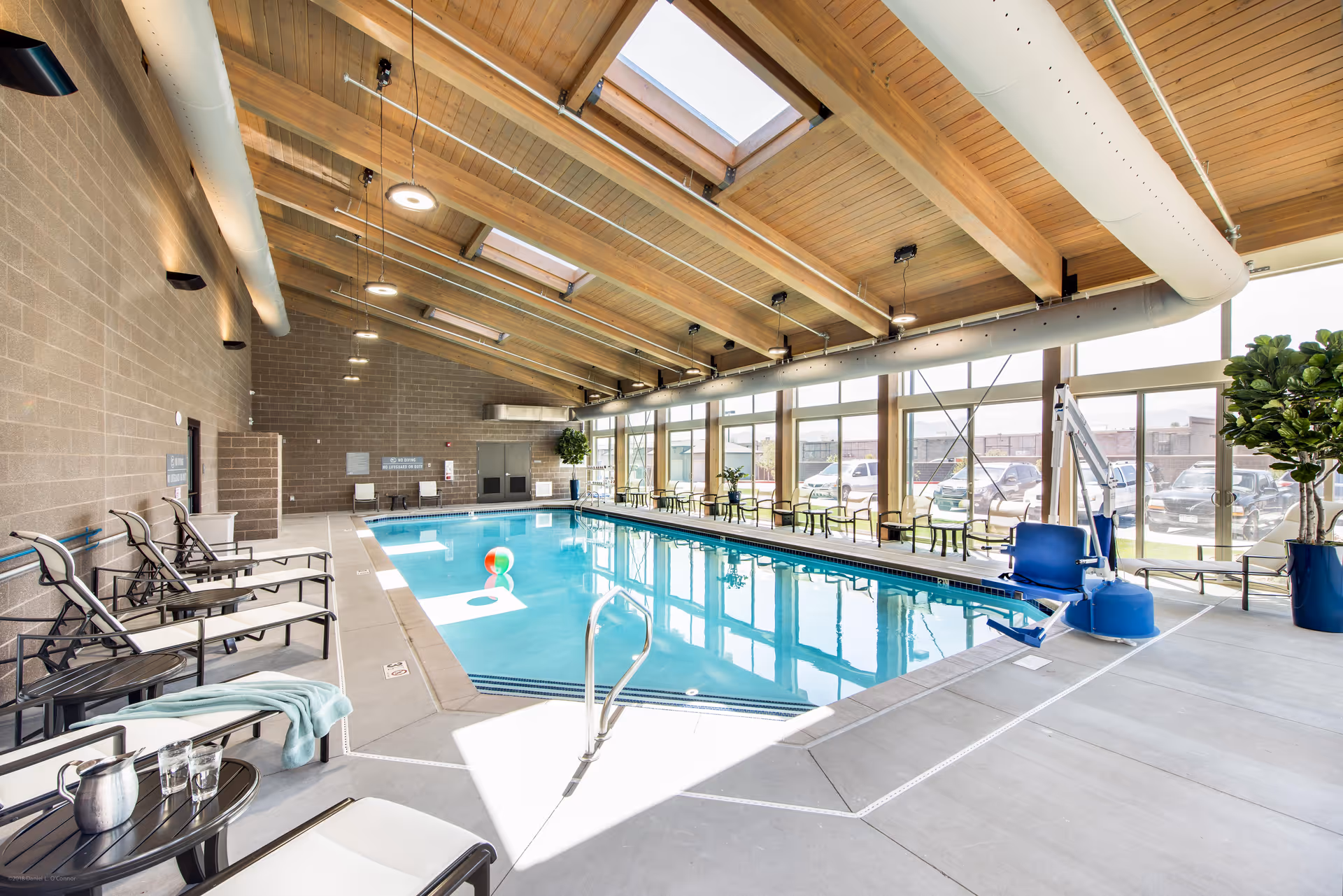 Indoor swimming pool area with lounge chairs, tables, and large windows letting in natural light. The ceiling has wooden beams and skylights, and there are plants placed around the pool area.
