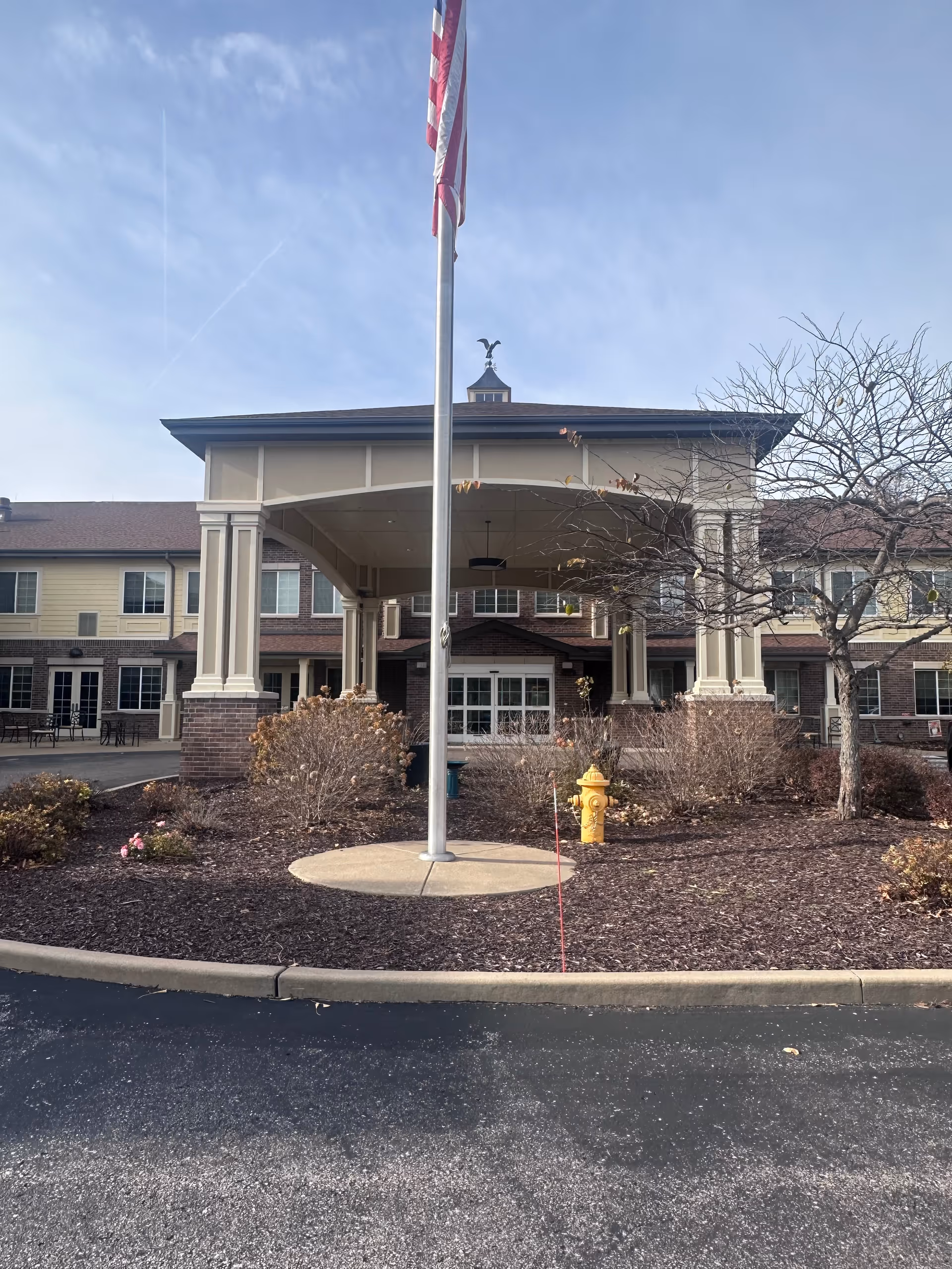 Front exterior view of Dougherty Ferry Assisted Living and Memory Care building with a covered entrance, an American flag on a flagpole, a yellow fire hydrant, and some leafless trees and shrubs in the landscaped area.