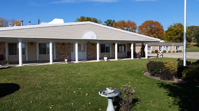 Single-story building with a covered porch supported by white columns, beige siding, and brick accents. The building is surrounded by a well-maintained green lawn with a birdbath and some bushes. Trees with autumn foliage are visible in the background under a clear blue sky.