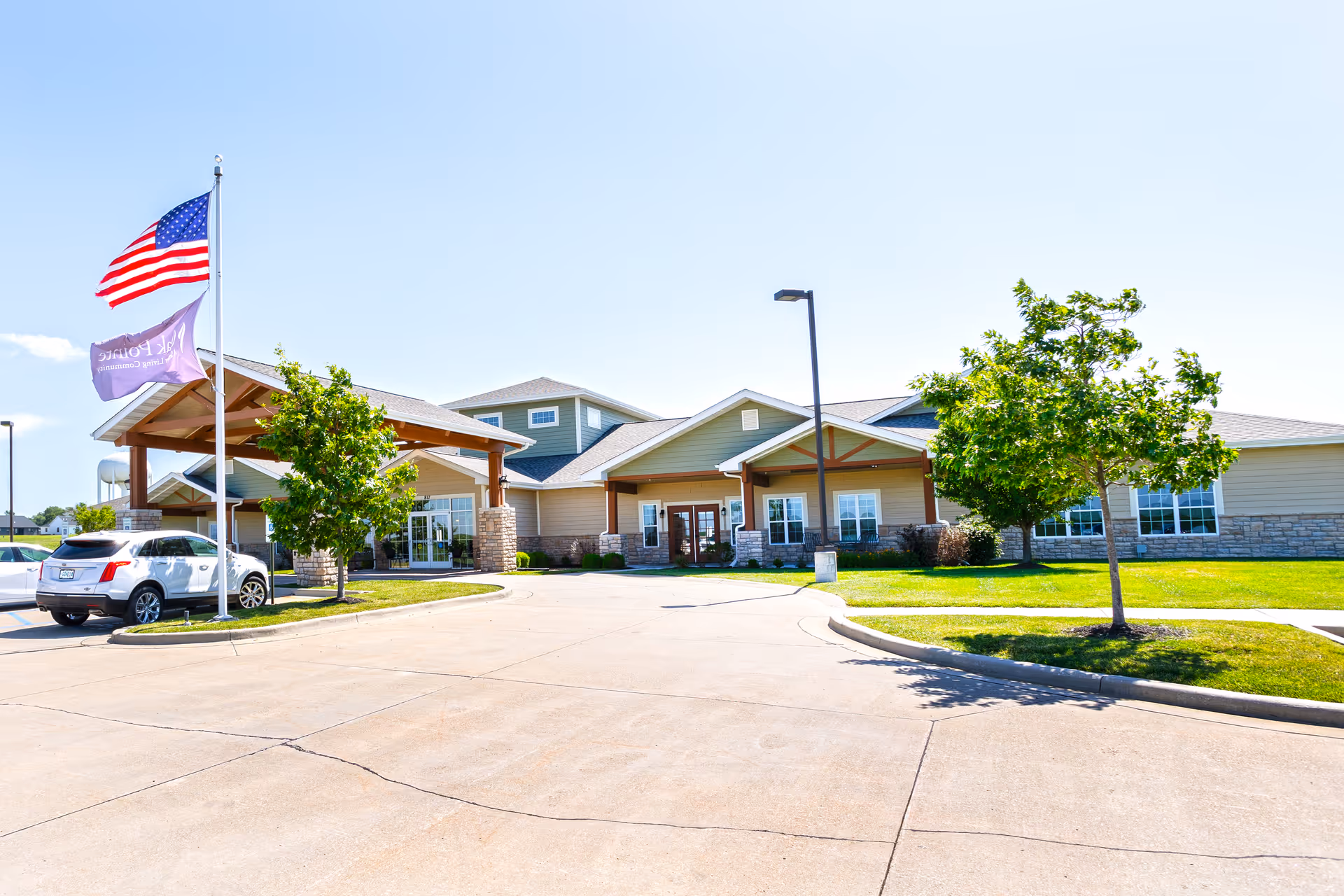 Front exterior of a senior living building with a covered entrance, flags, parked cars, and landscaped lawn.