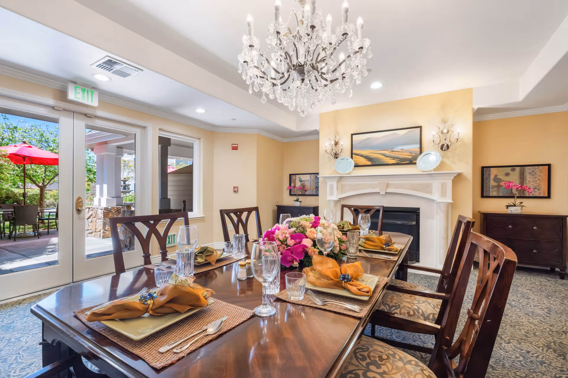 A well-decorated dining room with a polished wooden table set for six people, featuring plates, glasses, silverware, and orange napkins with napkin rings. A floral centerpiece with pink and purple flowers is in the middle of the table. The room has a chandelier hanging from the ceiling, a fireplace with decorative plates and a landscape painting above it, and large glass doors leading to an outdoor patio with red umbrellas and greenery.